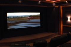 Home theater with a wide screen on a black wall, a raised stage and a wall sconce on wood trim
