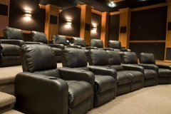 Tiered rows of black leather recliners in a home theater with wood columns and black fabric walls