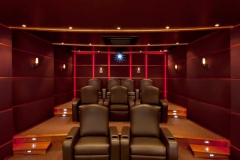Home theater seen from the screen: tiered leather recliners, lit steps, red LED accents and padded walls