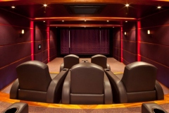 View from behind leather recliners toward a curtained screen, with red LED accents and padded walls