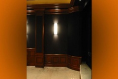 Corner of a theater with dark fabric walls, wood wainscot and a wall sconce, floor still bare