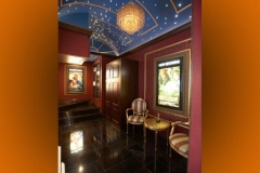 Theater lobby with a starry vaulted ceiling, chandelier, lit poster cases and a gilt chair and table