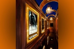 Corridor of marquee-lit poster frames under a blue barrel-vaulted ceiling with chandeliers