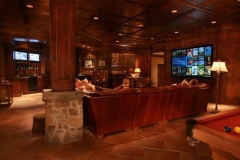 Media room with wood-paneled walls and ceiling, leather sectional facing a wall-mounted screen, bar and pool table