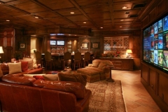 Media room with a large wall-mounted screen, leather seating on a rug, and a bar with stools under a coffered wood ceiling