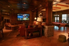 Media room with stone-clad wood columns, wall-mounted screen above a sofa, and a pool table by the windows